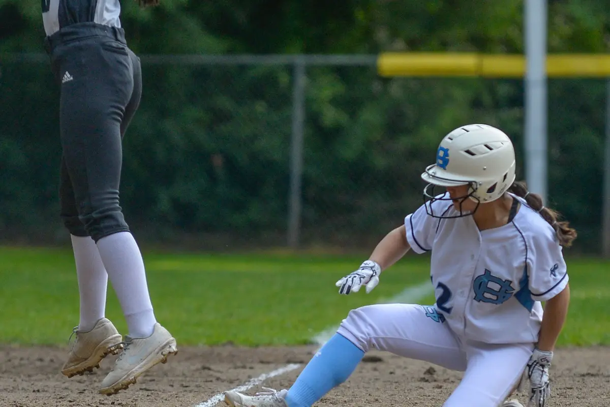 Softball player reaching to catch the ball as another player slides into base